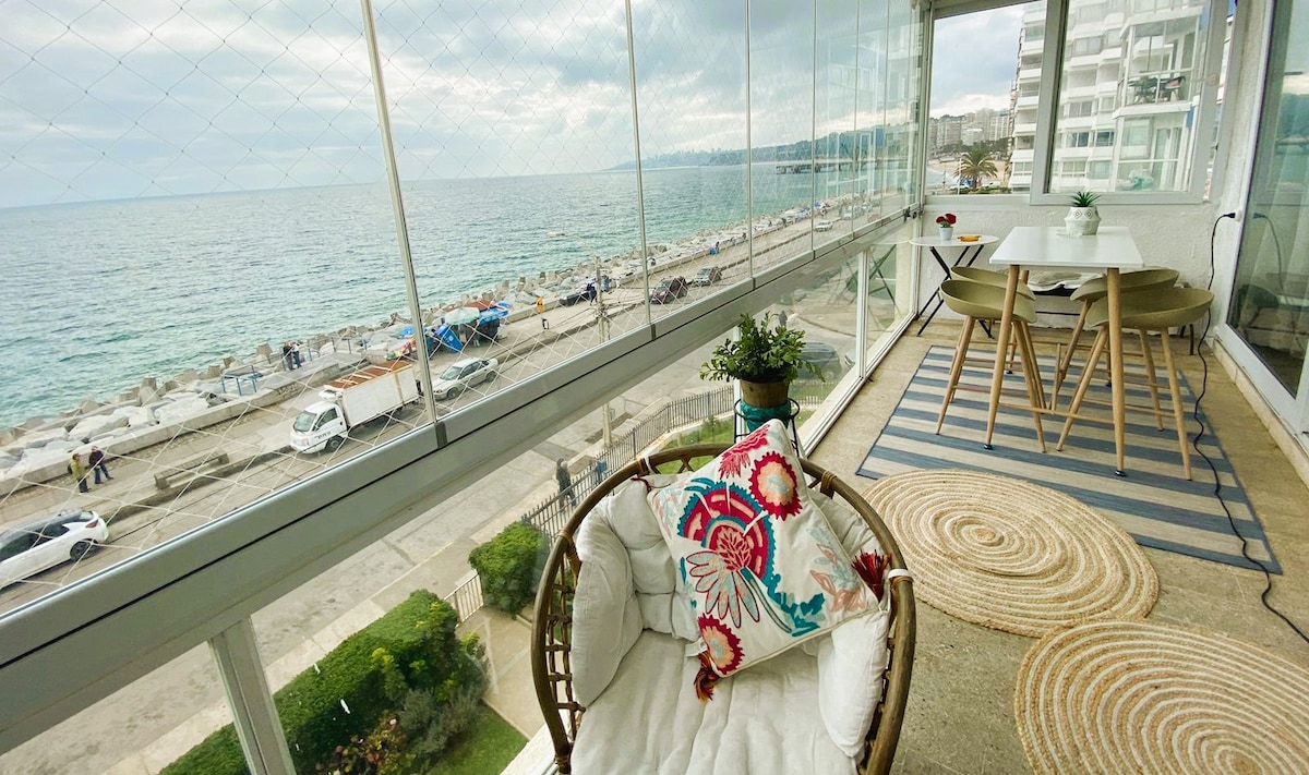 A sunlit balcony features a comfortable chair adorned with a decorative pillow, surrounded by clear glass railings that provide unobstructed views of the ocean. A small table with two chairs offers a cozy space for enjoying meals with serene beach scenery in the background.