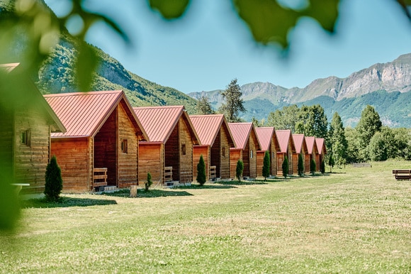 A row of eight wooden cabins features steeply pitched roofs and is set against a backdrop of mountains and clear blue skies. An expansive green lawn surrounds the cabins, adding to the natural ambiance of the area.