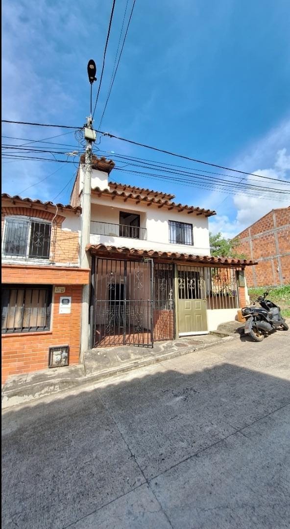 A two-story house with a traditional tile roof is showcased, featuring a gated entrance. The façade includes large windows and a covered porch area. Nearby, a motorcycle is parked along the side, with clear blue skies above.