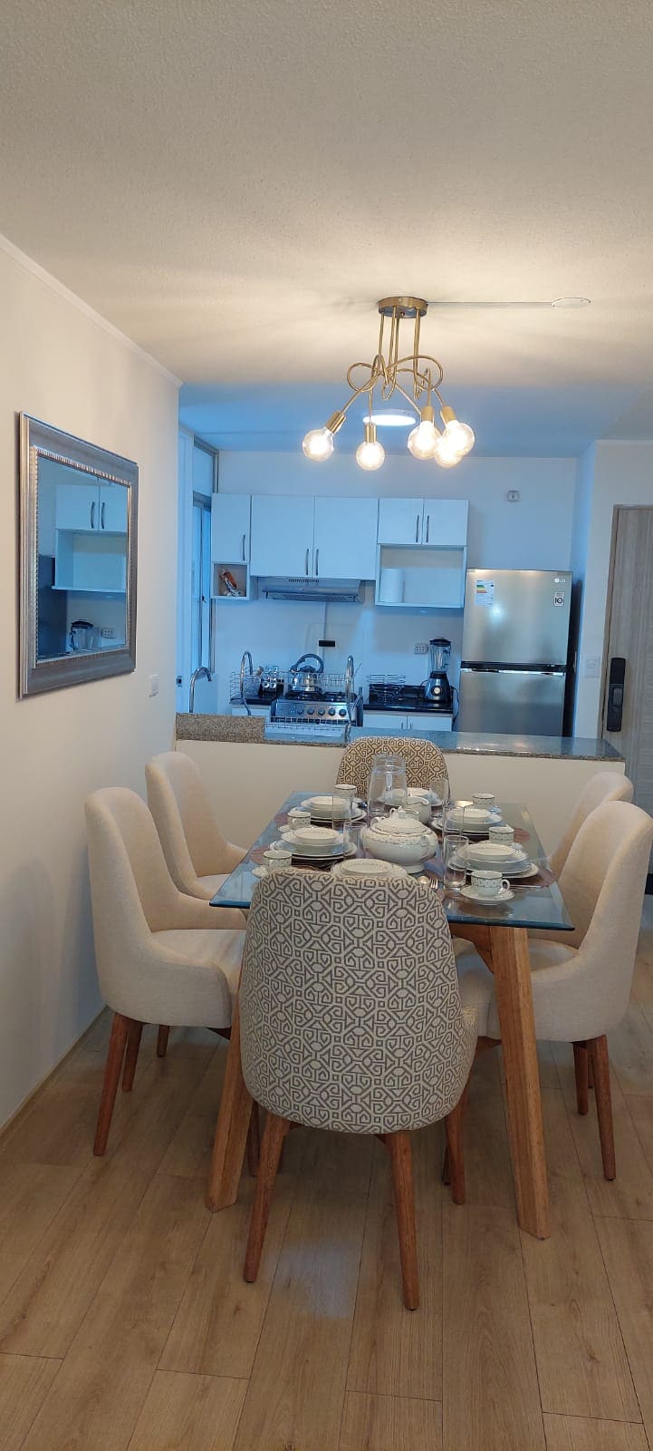 A dining area features a wooden table surrounded by four light-colored upholstered chairs. A mirror is mounted on the wall, reflecting the space. The kitchen is visible in the background, showcasing modern appliances, including a silver refrigerator.