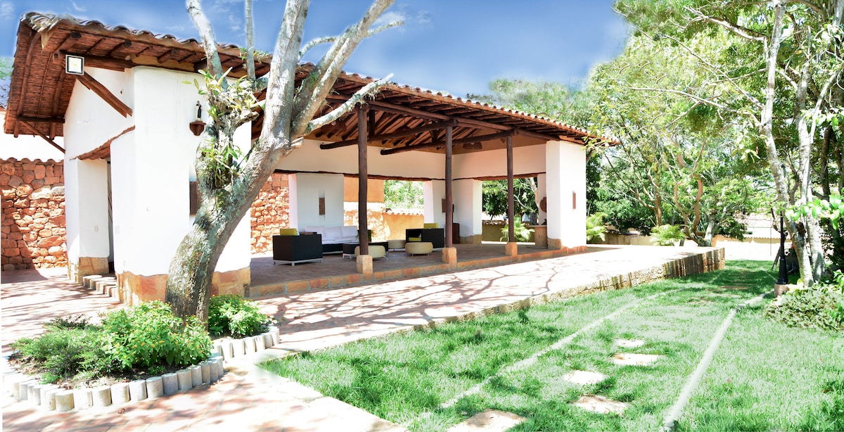 A spacious outdoor area is featured, with a covered seating space defined by wooden beams and a tiled roof. Comfortable seating arrangements are visible, surrounded by lush green grass and strategically placed trees, creating a serene environment for relaxation.