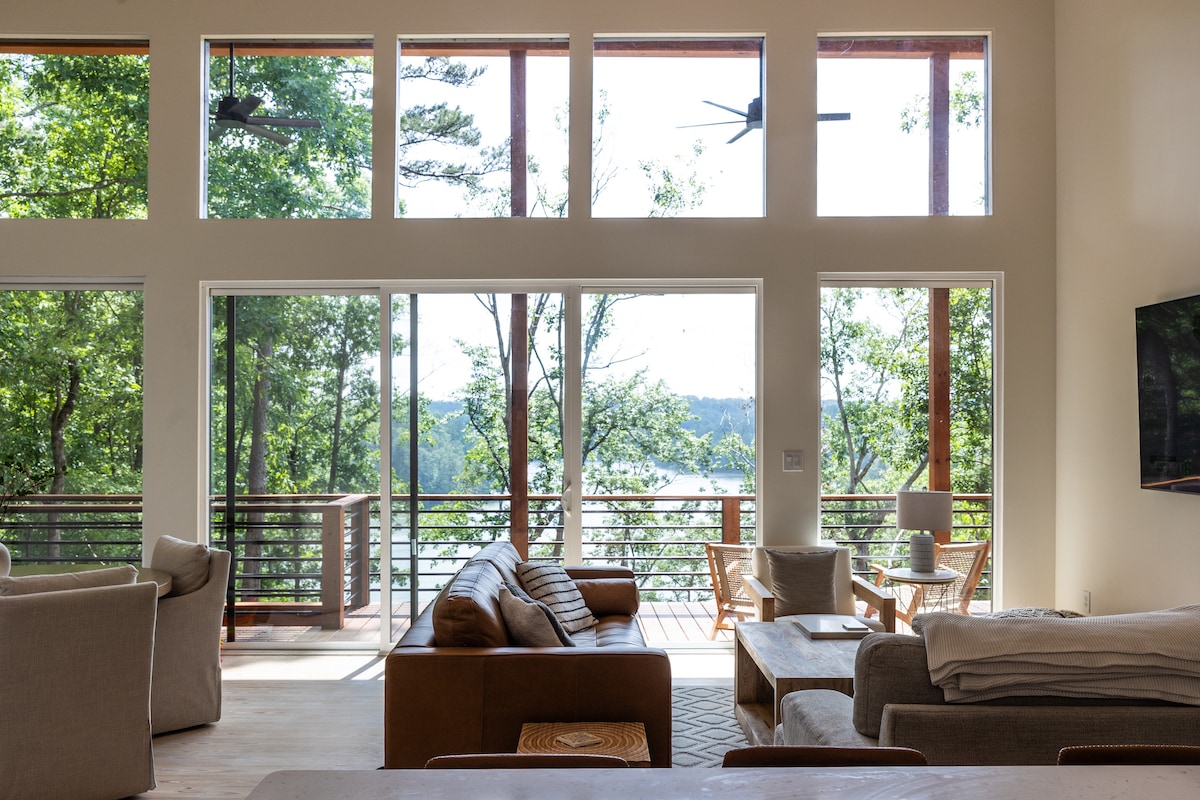 A spacious living area features large windows that fill the room with natural light, offering serene views of the surrounding trees and lake. Comfortable seating arrangements are visible, complemented by a cozy atmosphere created by neutral-toned furnishings.