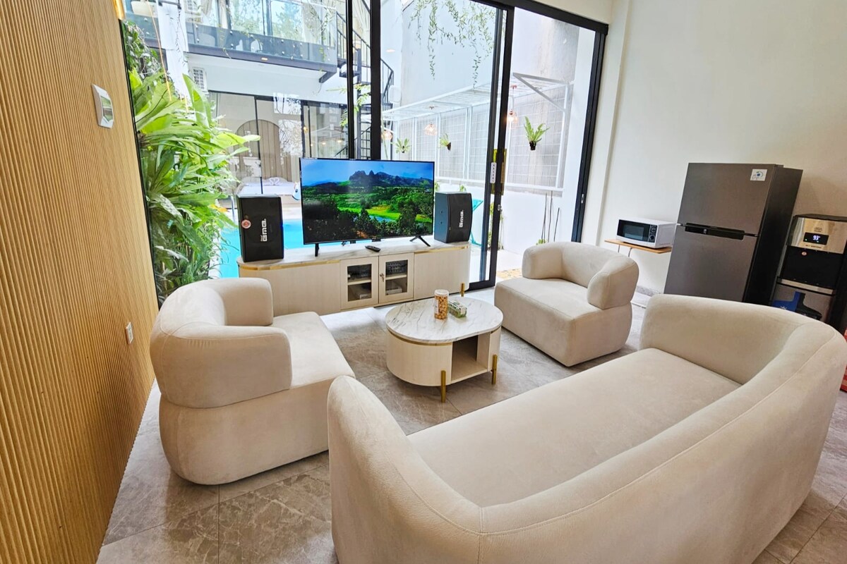 A modern living area features two soft, curved sofas positioned around a round coffee table. A large television displays a scenic view, while large windows provide ample natural light and glimpses of greenery outside. A sleek refrigerator is located nearby.