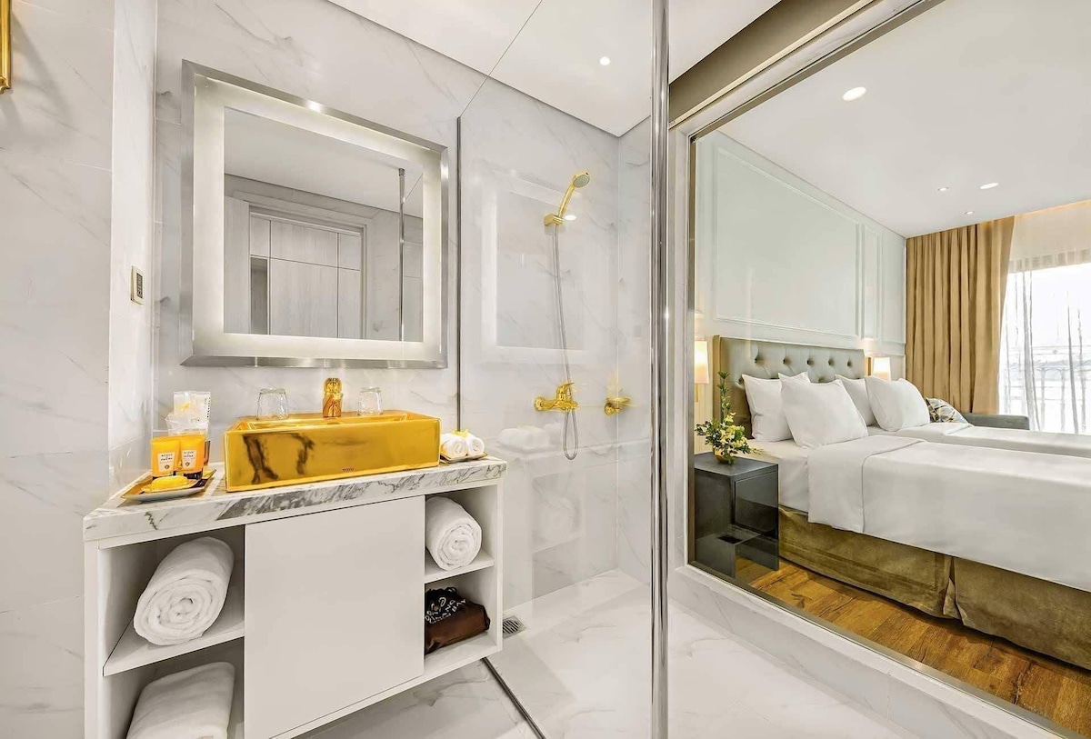A modern bathroom features a marble countertop with toiletries and neatly arranged towels. A glass shower is visible, with golden fixtures complementing a large mirror. The spacious bedroom is reflected in the mirror, showcasing a well-appointed setup with natural light from a nearby window.