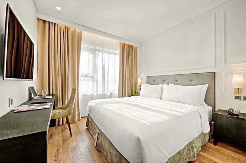 A well-appointed bedroom features a spacious bed dressed in crisp white linens. Natural light filters through large windows, illuminating the light-colored walls. A sleek desk and chair are positioned against one wall, accompanied by a flat-screen television mounted above a dark wood console.