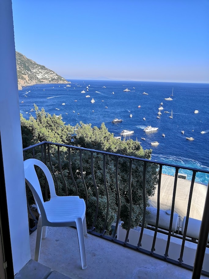A white plastic chair is positioned on a private terrace overlooking the vibrant blue sea dotted with boats. The coastline features lush greenery and rolling hills, creating a serene backdrop under a clear sky.