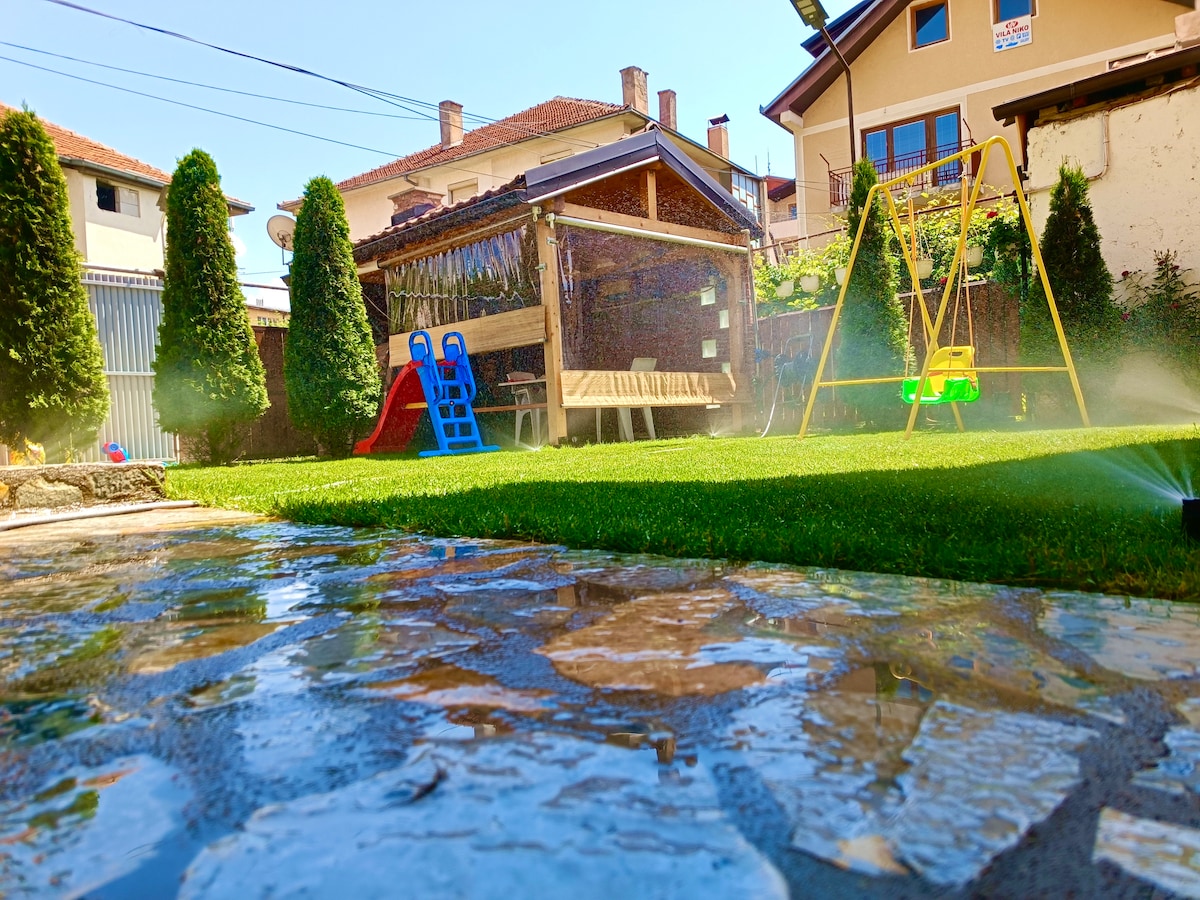 A well-maintained garden features manicured grass, a wooden swing set, and a colorful play structure. A stone pathway leads through the yard, with a wooden bench under a shaded pergola in the background. The surrounding trees provide a sense of privacy.