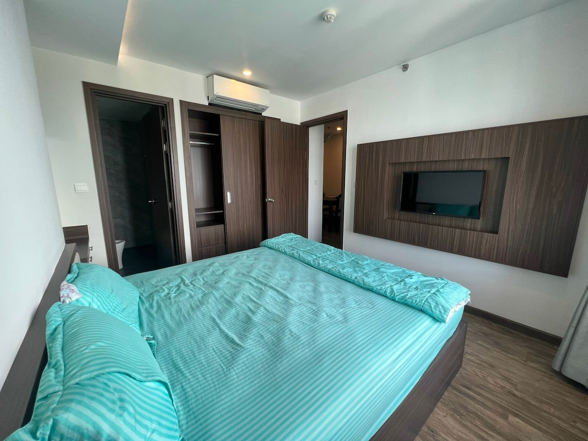 A spacious bedroom features a large arranged bed, covered with a soft teal bedding set. A mounted flat-screen television is displayed on the wall, and built-in closets are positioned nearby, providing ample storage space. Natural light enters through an unseen window.