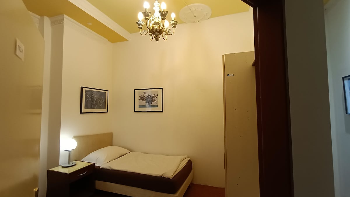 A simple room features a spacious bed with neutral bedding set against a light-colored wall. Two framed artworks are hung above the bed. A bedside table with a lamp and a small wardrobe are also visible. A chandelier provides soft lighting from the ceiling.