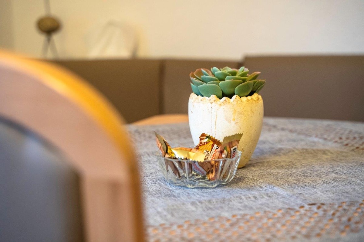 A dining table is set with a textured tablecloth. A small succulent plant is housed in a light-colored pot, while a crystal bowl filled with assorted chocolates rests nearby. Soft seating can be seen in the background.