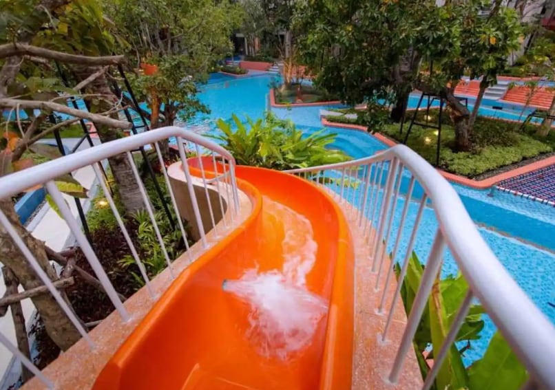 An orange water slide winds down alongside lush greenery, leading into a vibrant swimming pool area. Multiple pools are visible, surrounded by tropical plants and seating areas, creating an inviting space for relaxation and enjoyment.