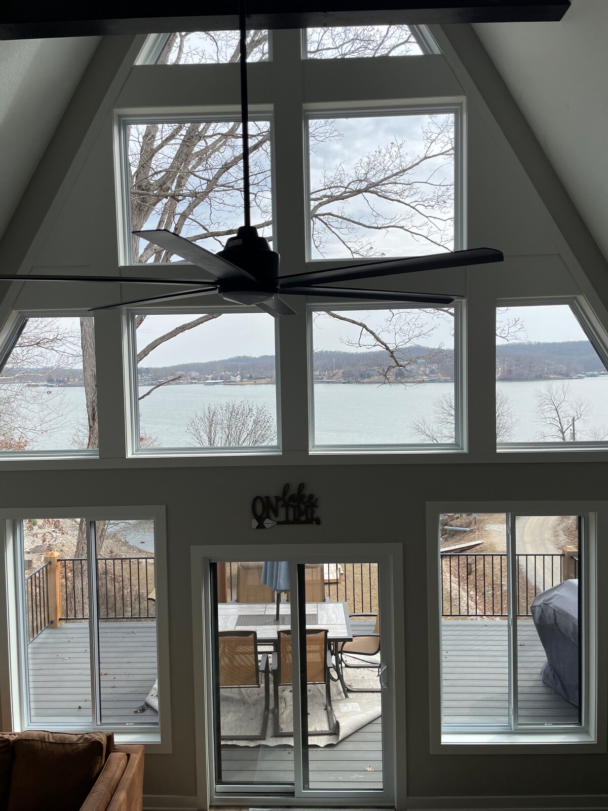 Large, floor-to-ceiling windows frame a view of the lake and surrounding trees. A ceiling fan hangs above, while a spacious outdoor deck with dining furniture is visible through sliding glass doors.