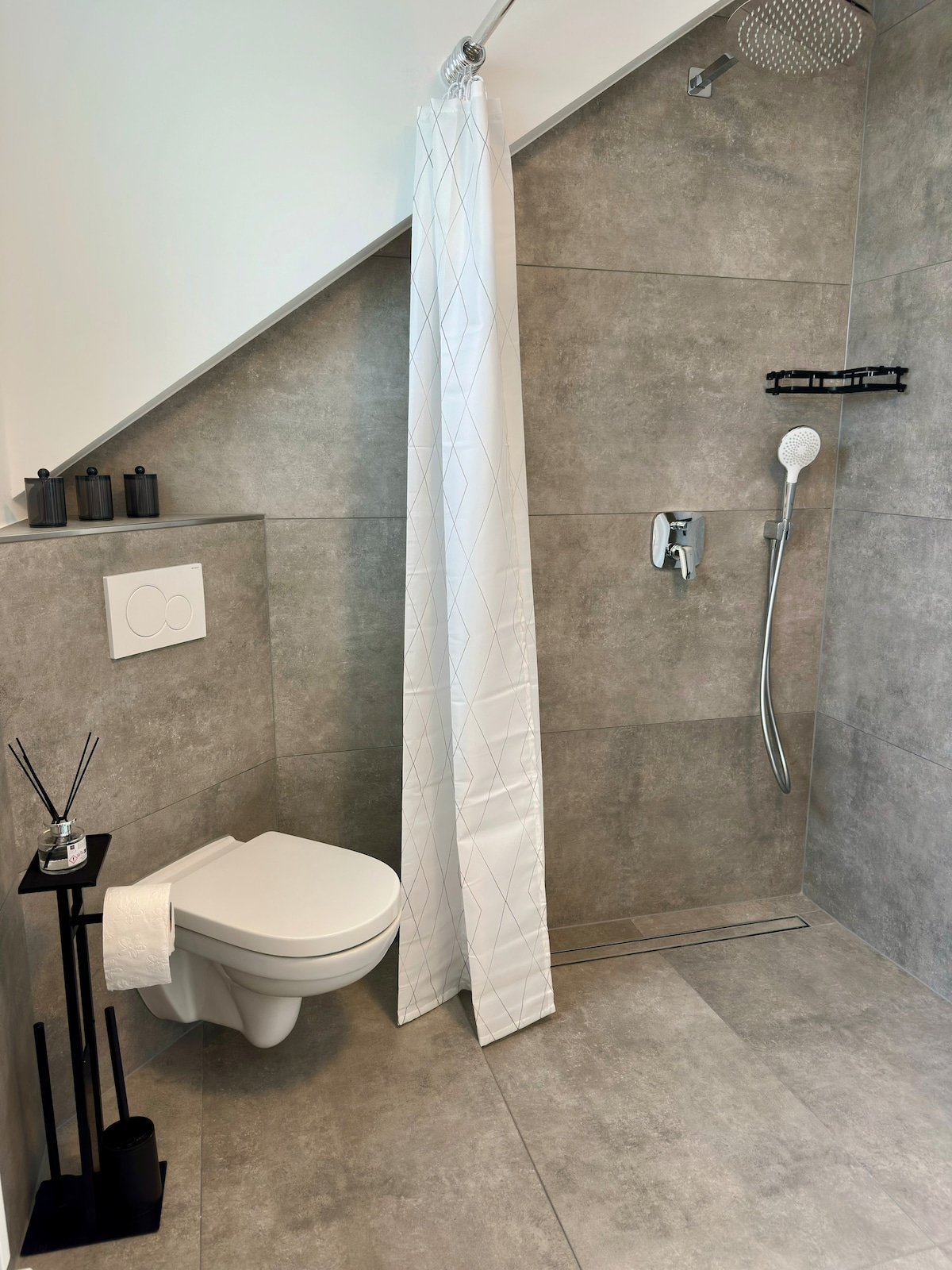 A modern bathroom features a sleek toilet and a shower area with a contemporary rainfall showerhead. Grey tiled walls and floors provide a uniform look, while a white shower curtain offers privacy. Decorative items are placed on a small black stand, enhancing the space's minimalist design.