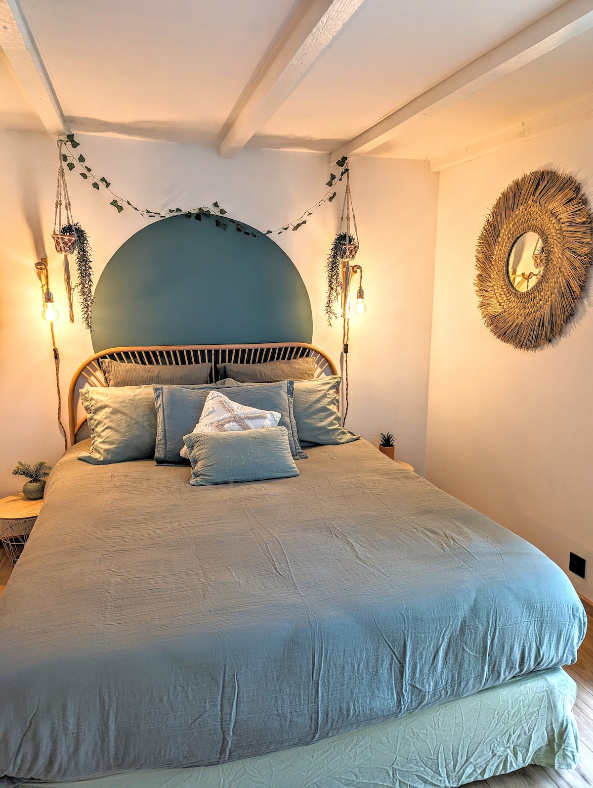 A cozy bedroom is featured with a spacious bed adorned with soft bedding and multiple decorative pillows. A soothing blue accent wall and natural lighting create a calming ambiance. Wall hangings and a round mirror add an elegant touch, enhancing the room's overall aesthetic.