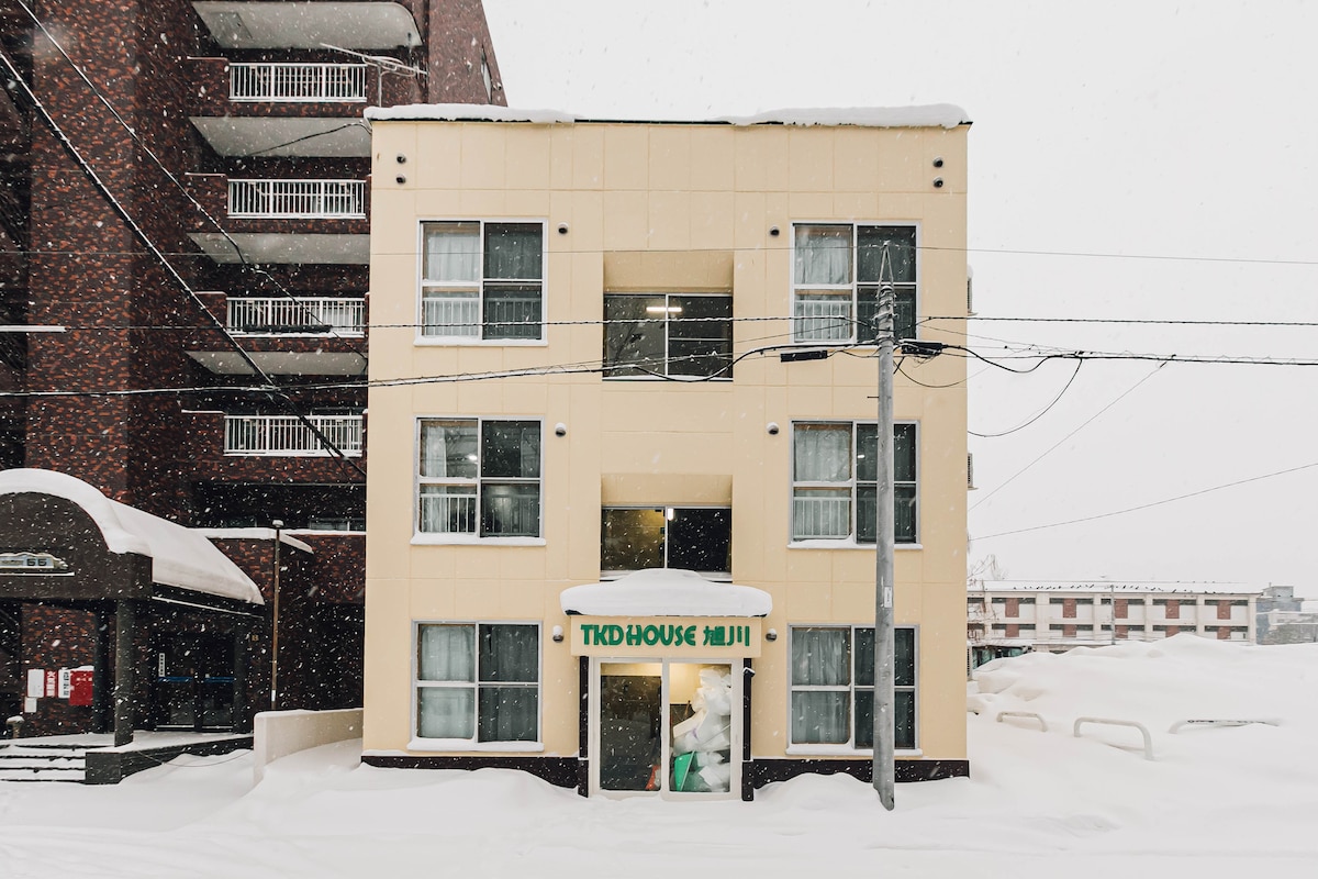 The exterior of TKD HOUSE旭川 is shown during winter, with snow blanketing the ground. The three-story building features multiple windows, and its entrance is marked by a green sign. Snow continues to fall gently, creating a serene, wintry atmosphere.