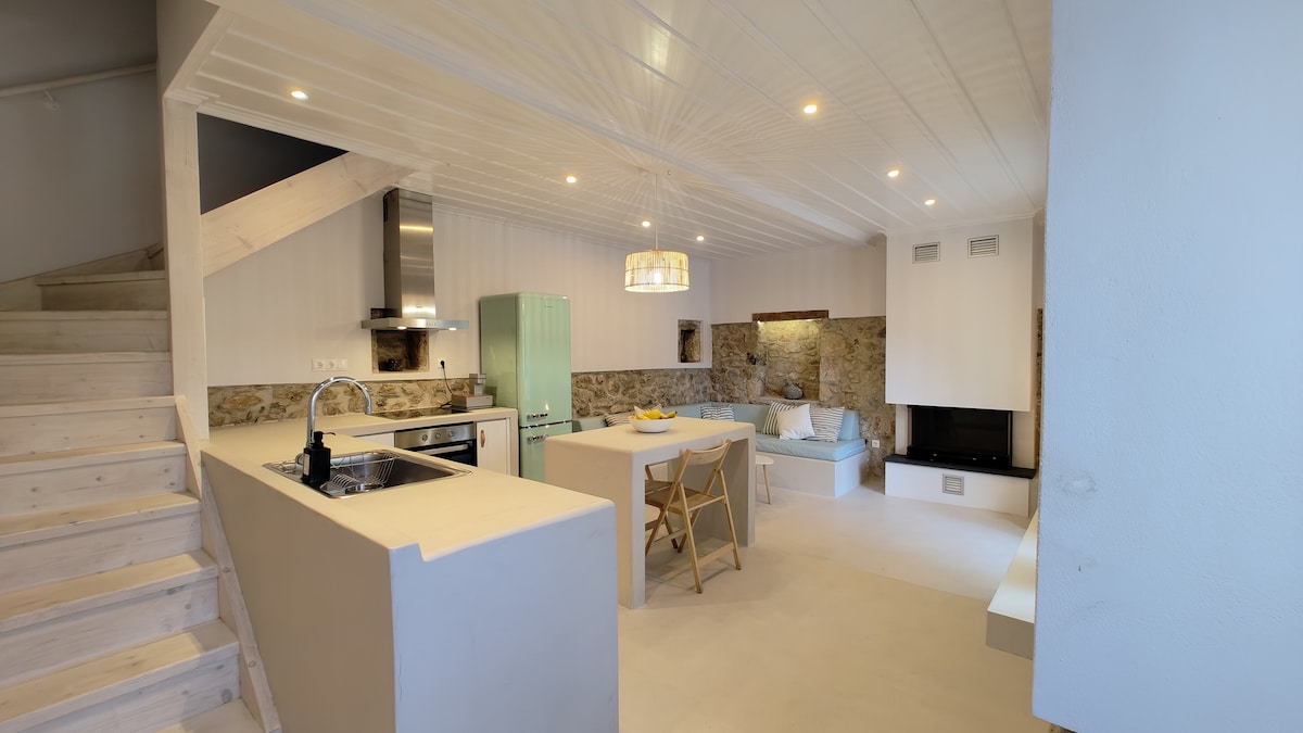 A bright and spacious kitchen-dining-living area is presented, featuring built-in counters and a central dining table-island. A comfortable corner sofa is located near an energy-efficient fireplace. Natural light enhances the clean design elements, with wooden accents visible throughout the space.