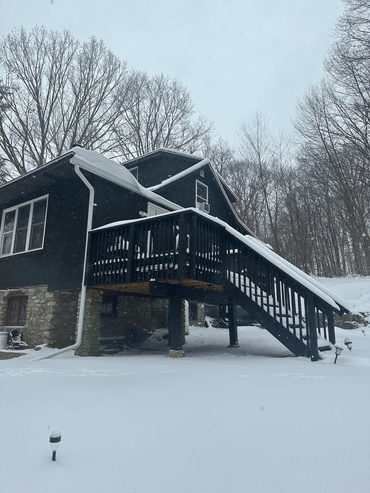 A charming home is depicted with a dark exterior and large windows, surrounded by a snowy landscape. A wooden staircase leads up to the entrance, while a blanket of fresh snow covers the ground, creating a serene winter atmosphere.
