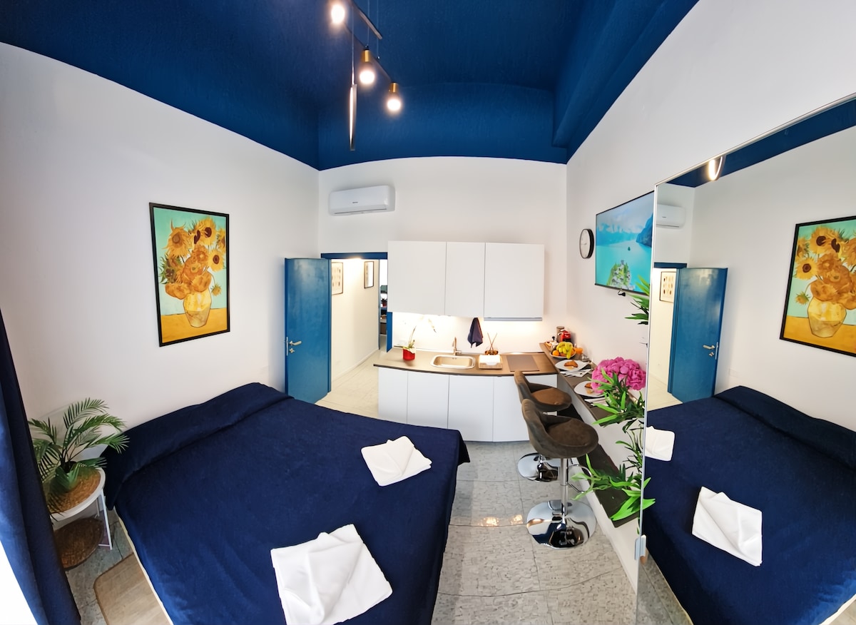 A bright room features a spacious bed with a blue coverlet, complemented by a decorative painting of sunflowers. A minimalist kitchen area is visible, along with a dining space and indoor plants. Light illuminates the room through two windows, enhancing the airy atmosphere.