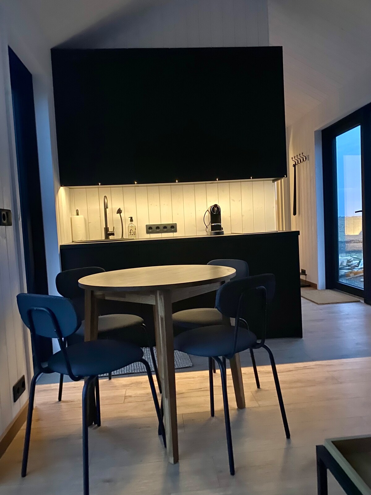 A compact kitchen area is showcased, featuring a dark cabinet with under-cabinet lighting. A round wooden table is accompanied by four minimalist chairs. Essential appliances, including an induction cooktop and a Nespresso coffee maker, are visible on the countertop.