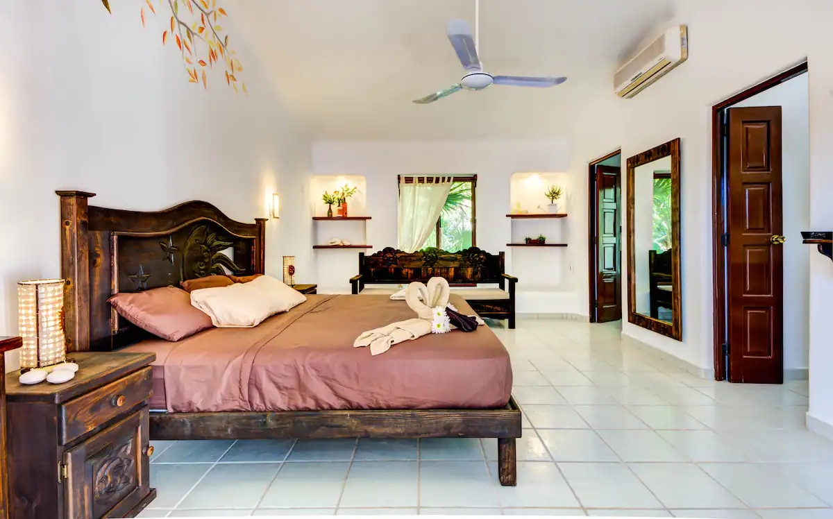 A spacious bedroom features a wooden king-size bed with warm-toned bedding. Two nightstands are positioned on either side. Natural light filters through sheer curtains, highlighting the tiled floor. There are decorative elements, a comfortable seating area, and air conditioning unit installed on the wall.