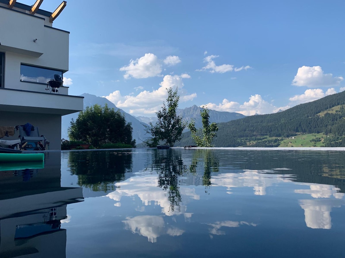 An infinity pool reflects clouds and mountains, creating a serene view. The pool's edge aligns with the landscape, and greenery is visible nearby, enhancing the outdoor setting. The modern building's structure is partially visible, suggesting a well-integrated design with nature.