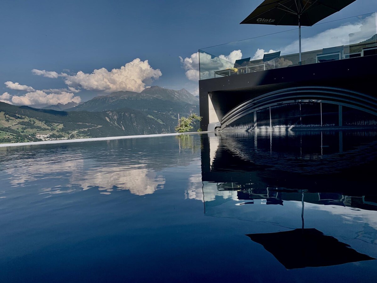 An infinity pool reflects the clear blue sky and surrounding mountains. Sun loungers are positioned beside the pool, shaded by umbrellas. The serene landscape creates a tranquil backdrop, enhancing the outdoor experience.