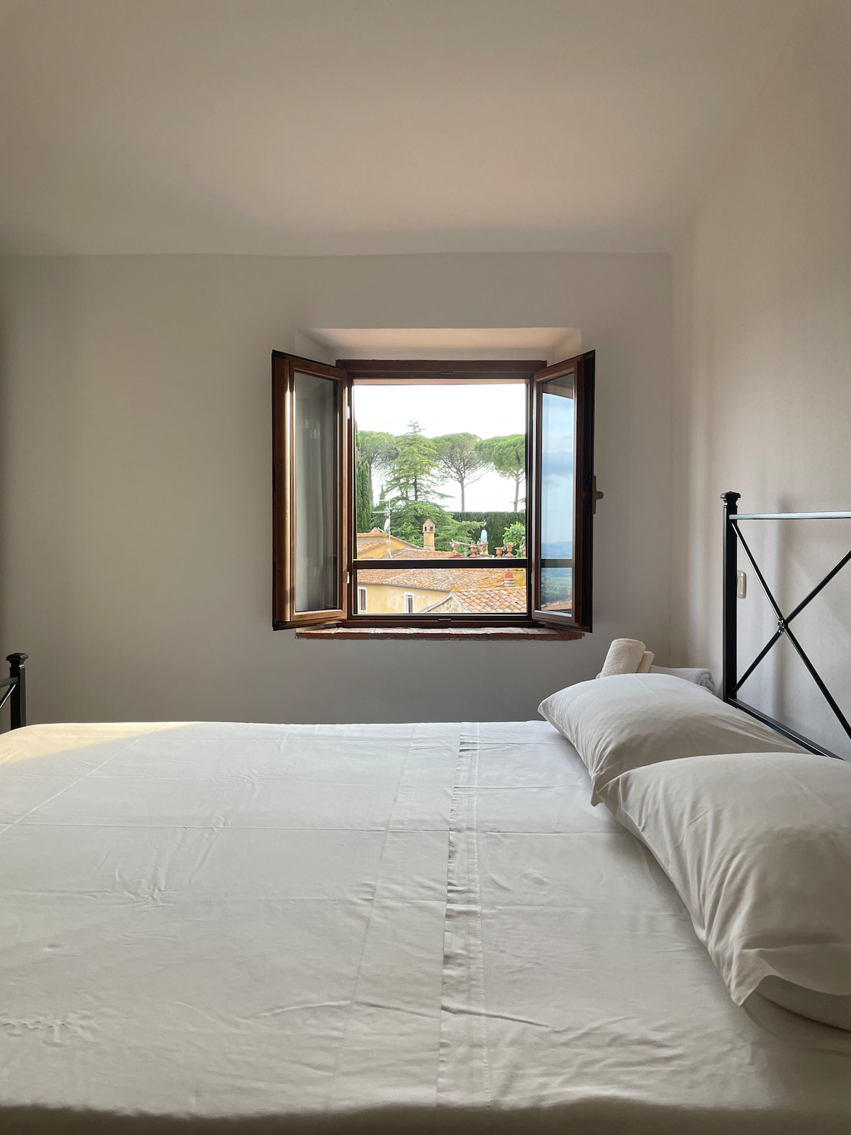 A bright bedroom features a spacious bed with crisp white linens. An open window allows fresh air and natural light to enter, revealing a view of nearby trees and rooftops. The room is painted in soft, neutral tones, contributing to a serene ambiance.