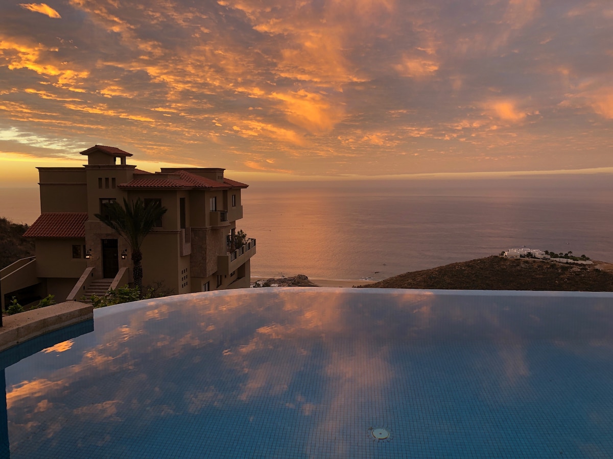 A tranquil scene captures the serene infinity pool reflecting vibrant sunset hues. The ocean stretches beyond, with a distant coastal view, while a beautifully designed villa stands to the side, framed by gentle clouds and the warm glow of twilight.