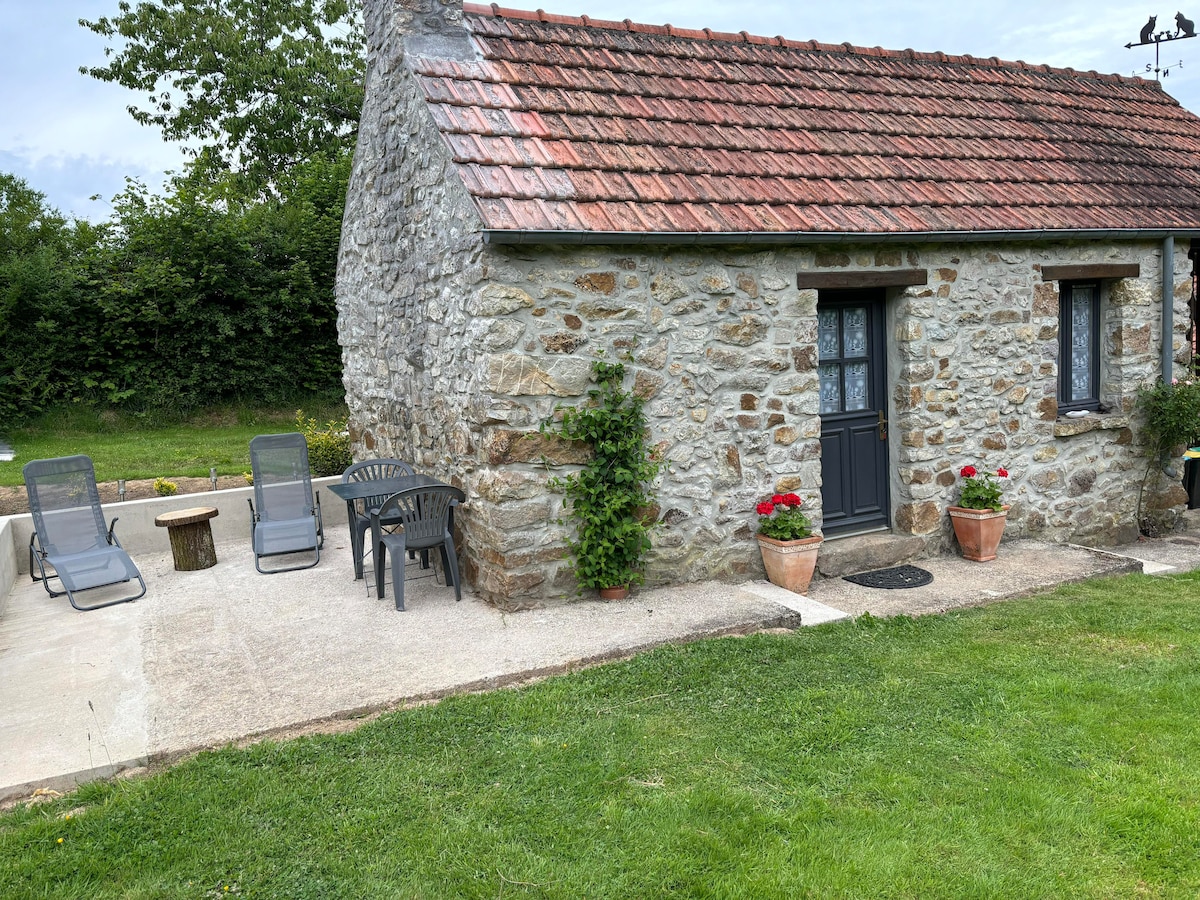 The exterior of a rustic stone cottage is featured, showcasing a sloped, tiled roof. A patio area includes two lounge chairs and a small table surrounded by flower pots. Lush green grass surrounds the cottage, enhancing the serene countryside setting.
