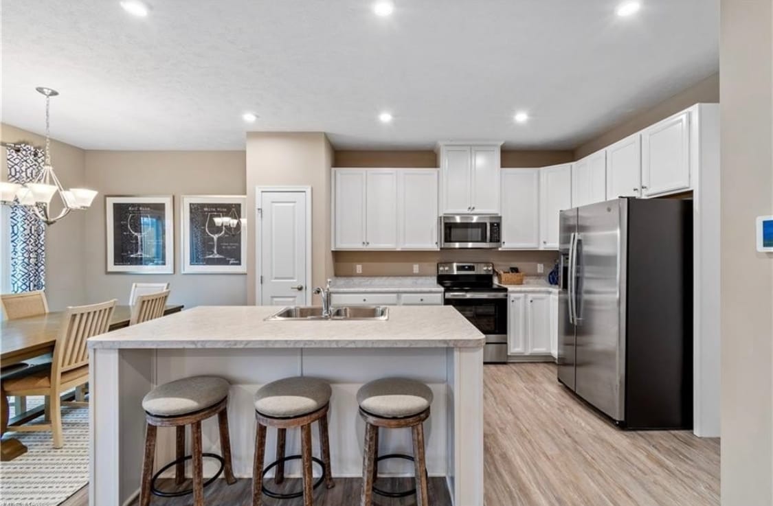 A bright kitchen with white cabinetry and stainless steel appliances is featured. An island with seating provides a central gathering space. A dining area with a wooden table and chairs is positioned nearby, complemented by framed artwork on the walls.