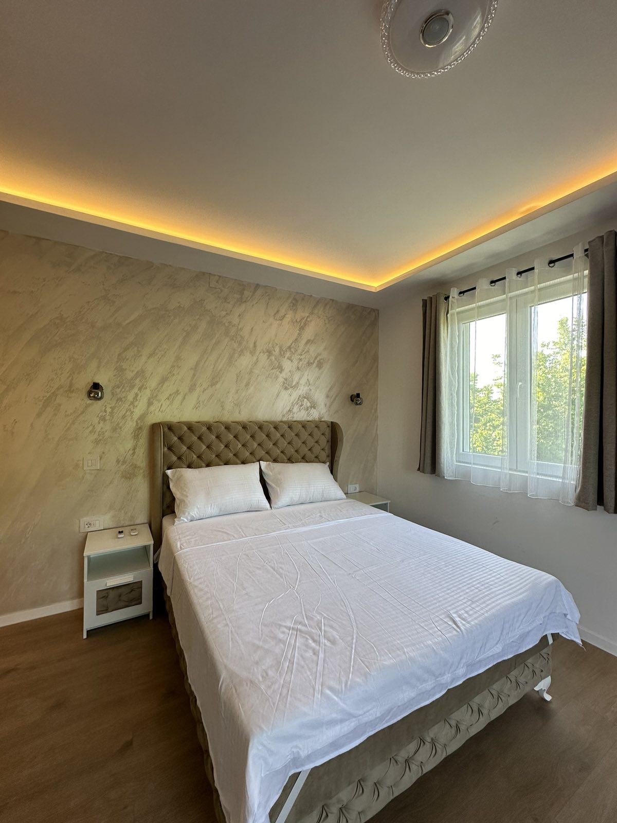 A light-filled sleeping area features a spacious bed with a tufted headboard and fresh white linens. The softly illuminated ceiling adds a warm glow, while a nearby window is adorned with light curtains, allowing natural light to enter the room.