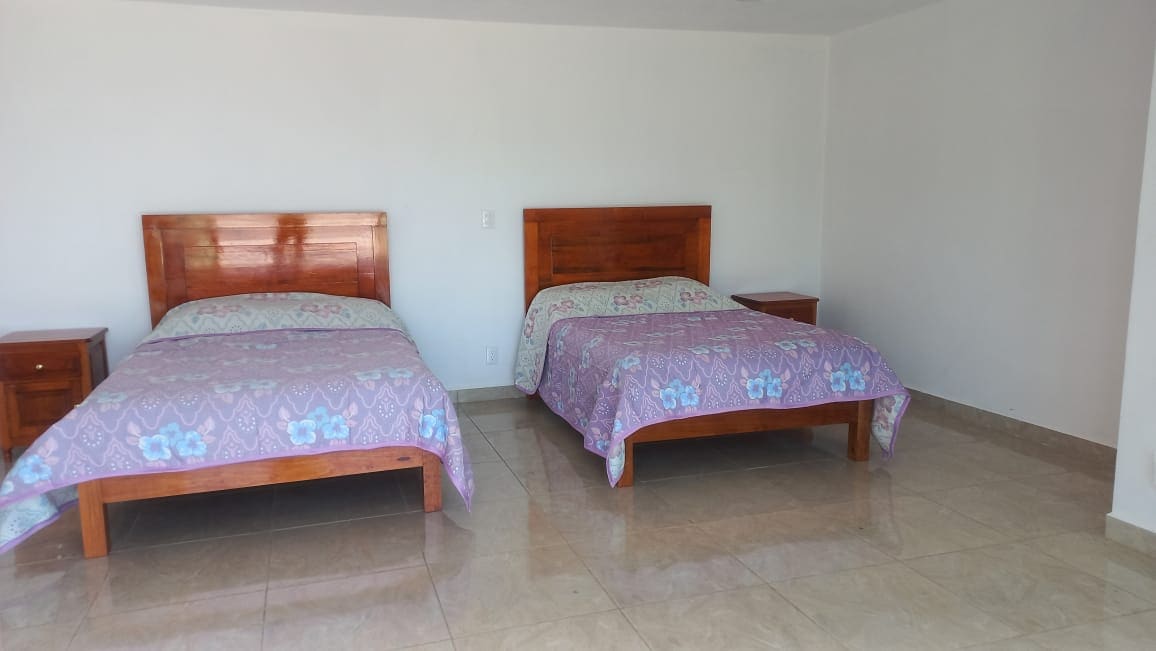 A spacious sleeping area features two wooden beds covered with patterned blankets. Nightstands are positioned beside each bed. The room is well-lit by natural light reflecting off the glossy tile floor, enhancing the simplicity of the white walls.