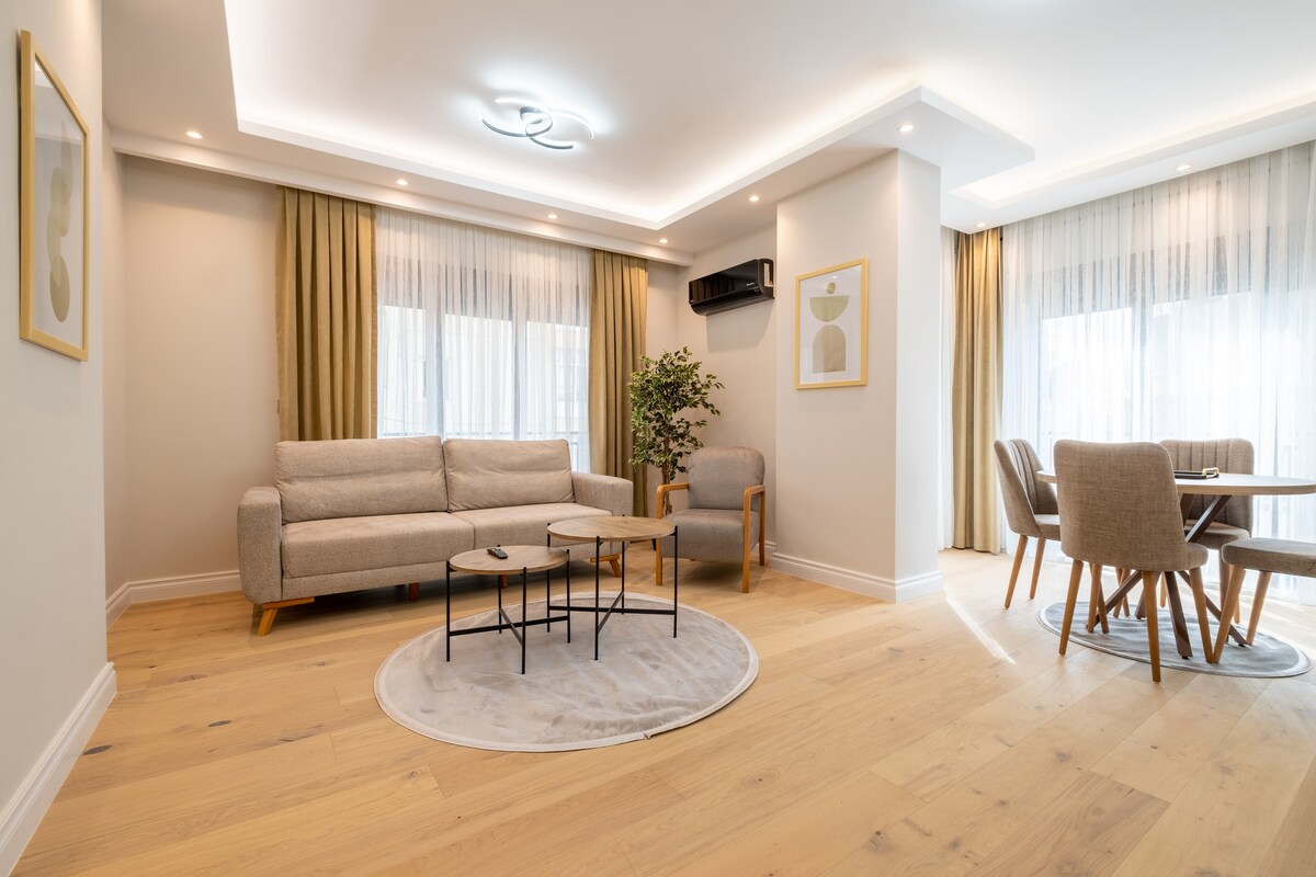 A spacious living area features two seating arrangements, including a light grey sofa and armchair. A circular area rug is positioned under a coffee table. Large windows with sheer curtains allow natural light to fill the room. A dining table with several chairs is situated nearby.