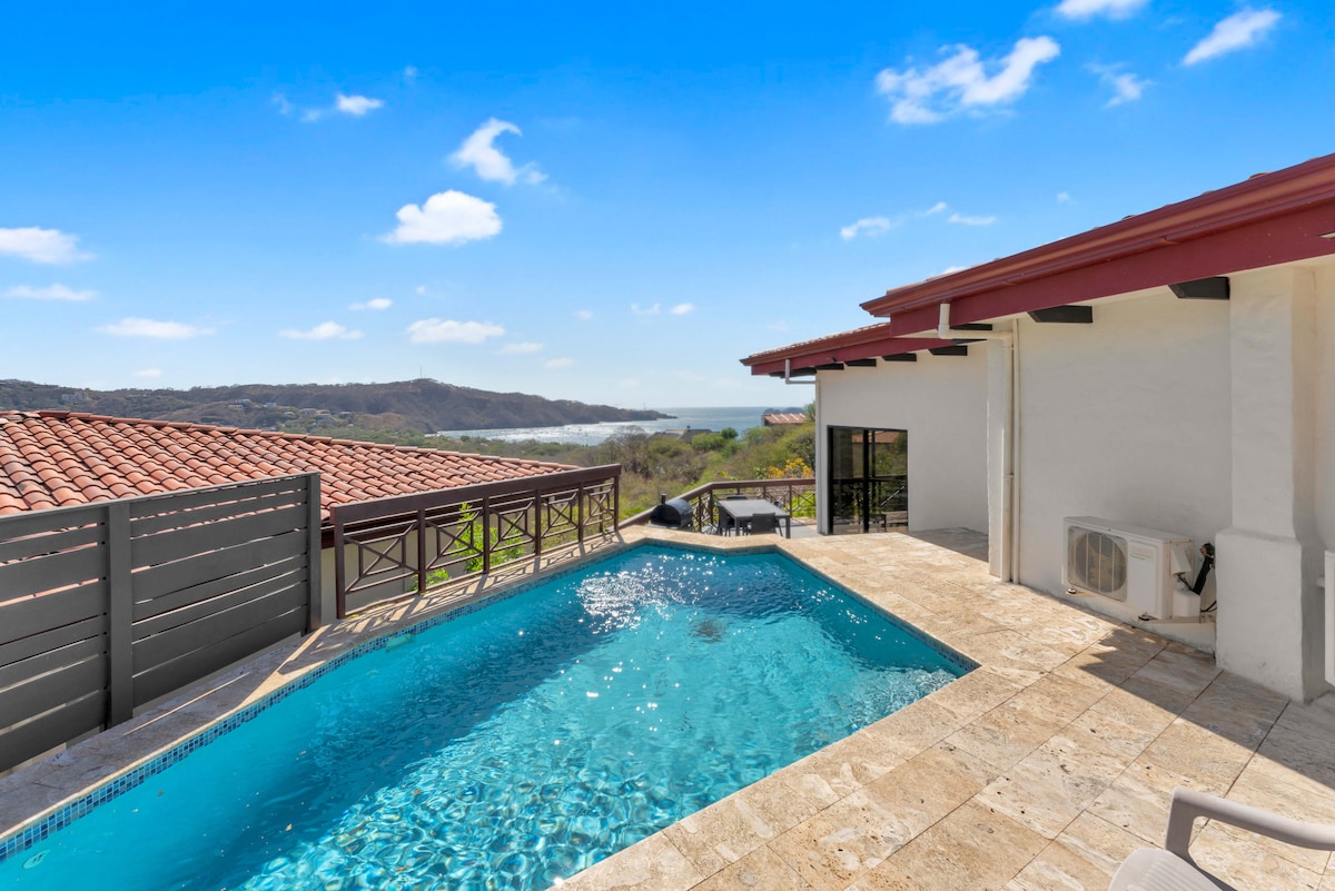 A private pool glistens under the sun, framed by elegant stone tiles. The expansive outdoor space offers views of the surrounding hillside and distant coastline, with clear skies and soft clouds creating a serene atmosphere. Large sliding doors provide access to the indoor areas.