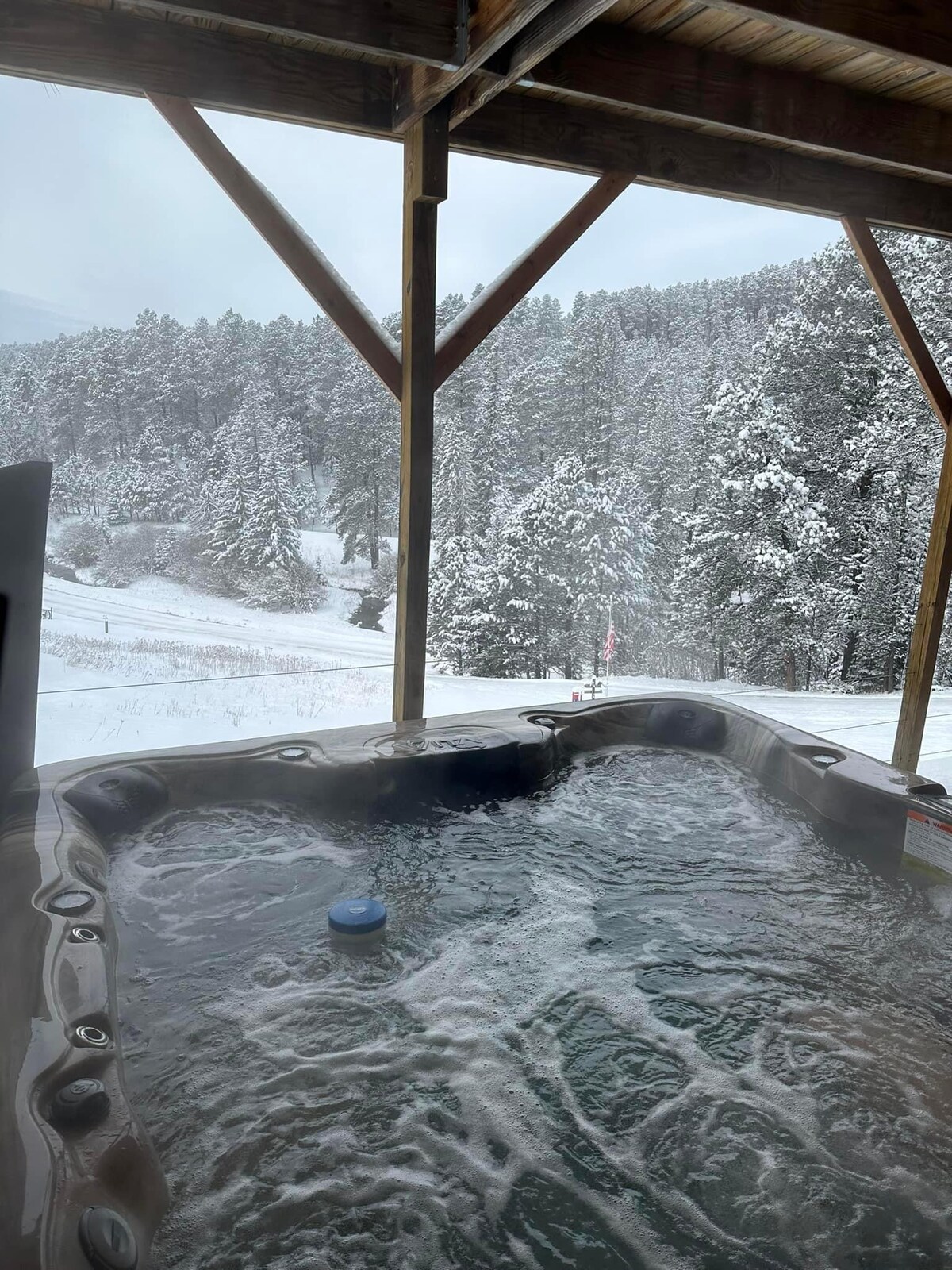 A hot tub is situated under a wooden structure, offering views of a snowy landscape filled with evergreen trees. Soft steam rises from the bubbling water, contrasting against the cold environment, creating a serene outdoor relaxation area.