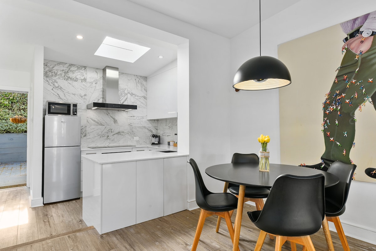 A modern kitchen area features sleek white cabinetry and a marble backsplash. A round dining table with black chairs is complemented by fresh flowers. Natural light floods the space, highlighted by a skylight. A refrigerator and stylish artwork are visible in the background.