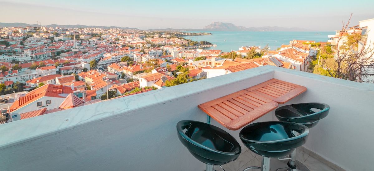 A balcony is featured, offering a panoramic view of the vibrant cityscape and ocean. Three sleek black bar stools surround a wooden table, inviting relaxation while enjoying the scenery. The horizon showcases a blend of rooftops and natural landscapes under a clear sky.