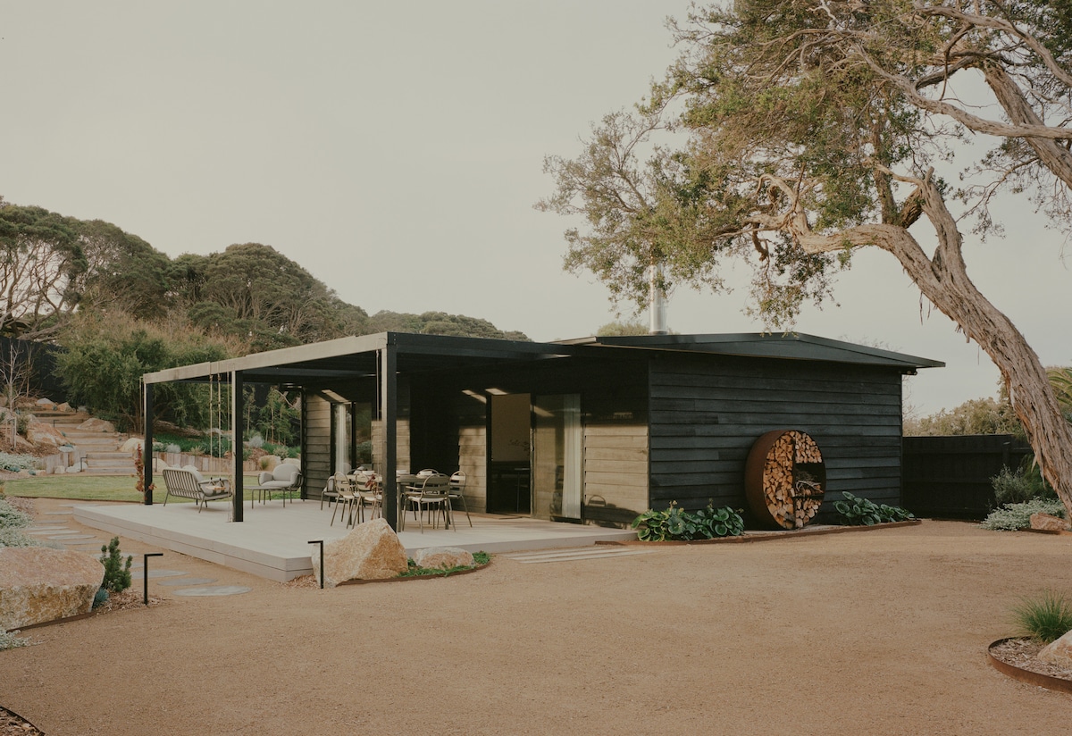 A modern single-story home features a sleek black exterior and expansive outdoor decking. A spacious dining area is visible, accompanied by a well-manicured garden, natural stone accents, and large trees, creating a harmonious blend with the surrounding landscape.