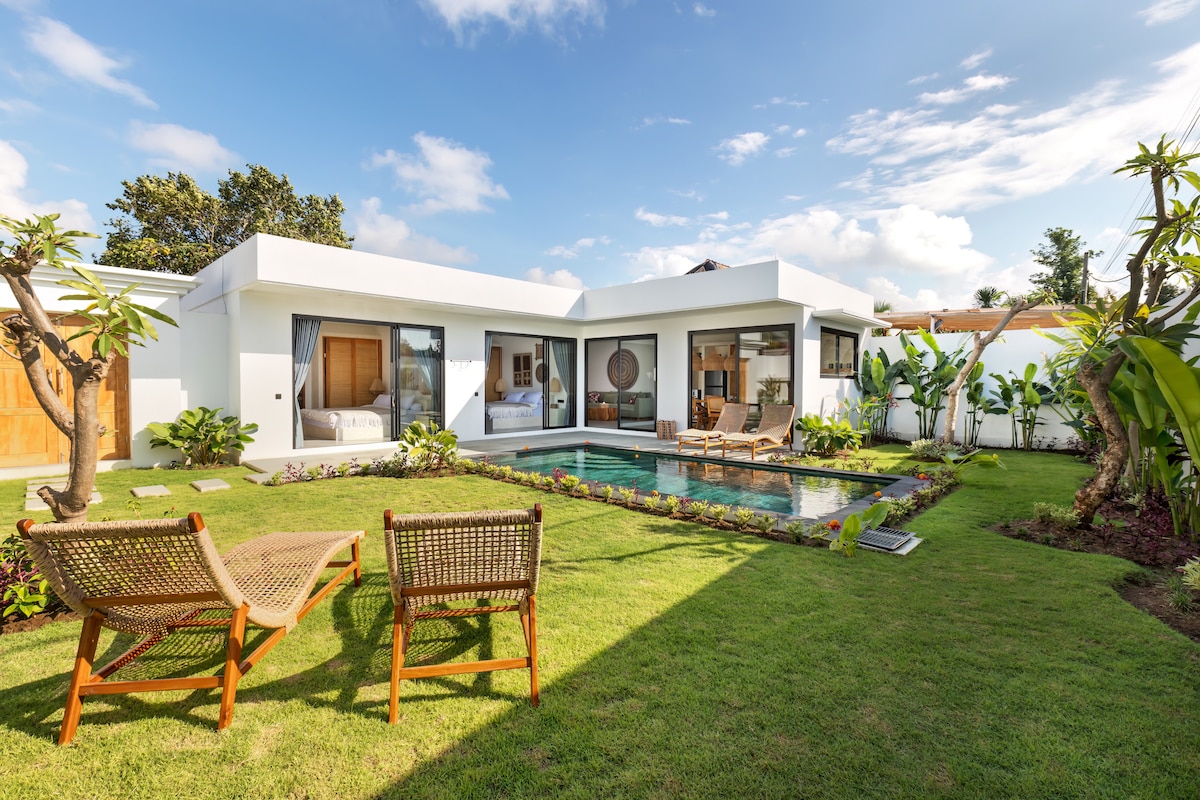 A modern villa is surrounded by a lush tropical garden, featuring a private swimming pool at its center. Comfortable outdoor chairs are positioned on the lawn, offering a perfect view of the villa's spacious interior through large glass doors. Greenery adds a serene touch to the setting.