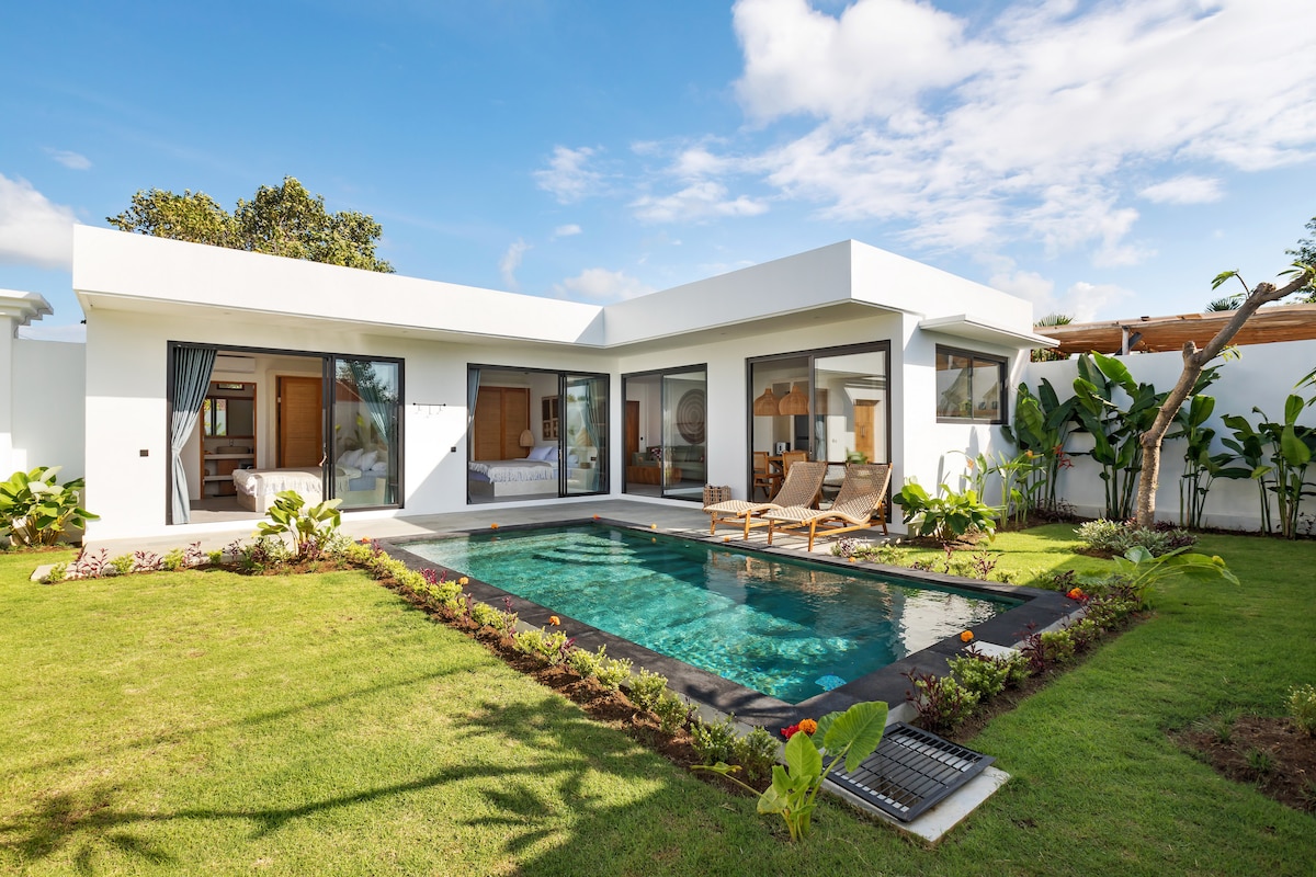 A modern villa is set within a lush green garden, featuring a private swimming pool surrounded by comfortable lounge chairs. Large glass doors open to the interior, creating a bright and inviting atmosphere with tropical plants framing the landscape.