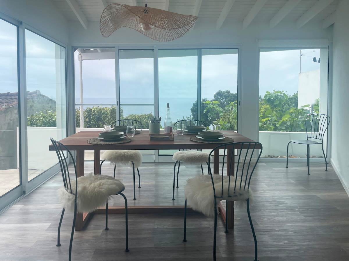 A dining area features a wooden table surrounded by four chairs, each adorned with soft covers for comfort. Large glass doors allow natural light to illuminate the space, offering views of the ocean and greenery beyond.