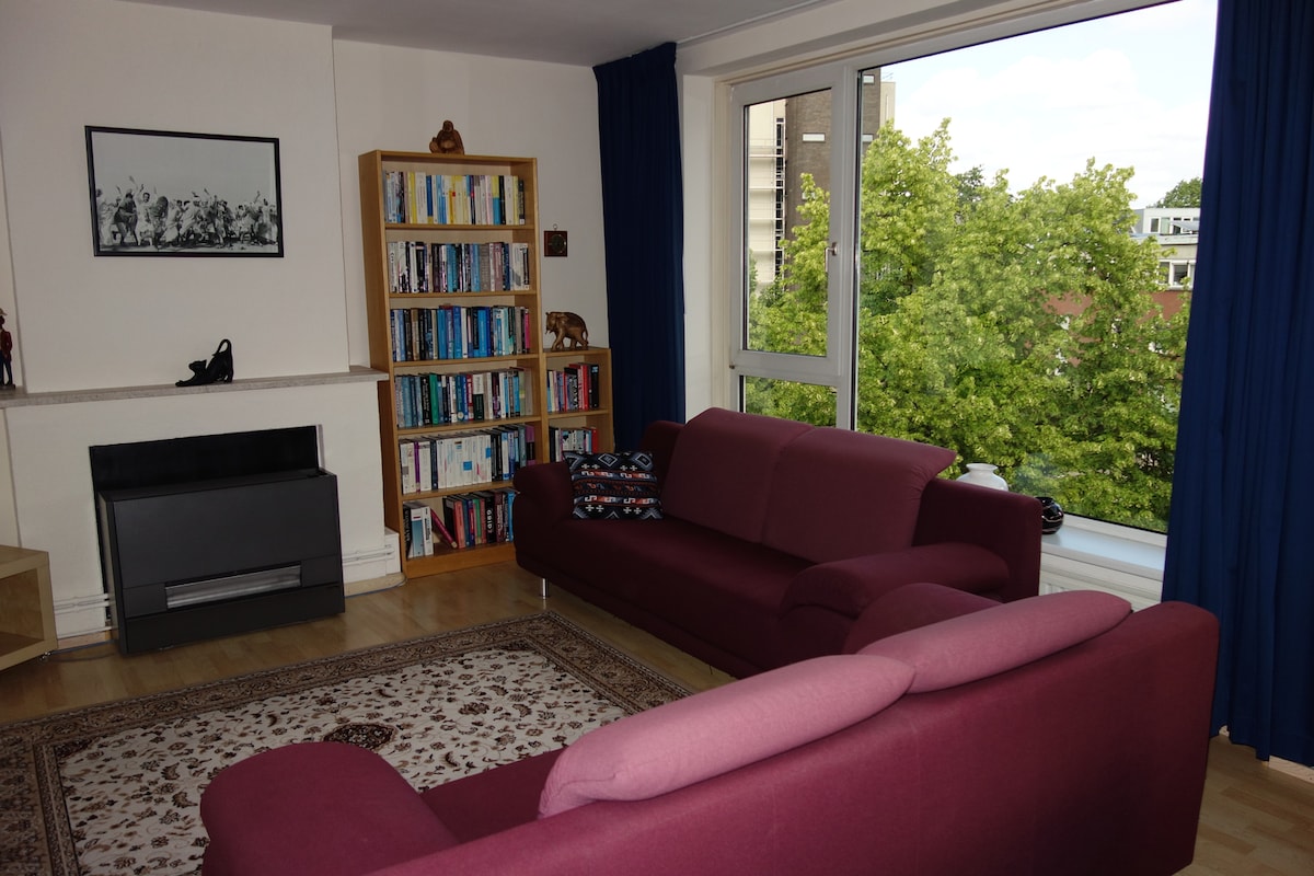 A well-lit living room features two sofas arranged around a decorative rug. A bookshelf filled with various books is positioned against the wall, while a large window allows natural light and offers views of greenery outside. Dark curtains frame the window.