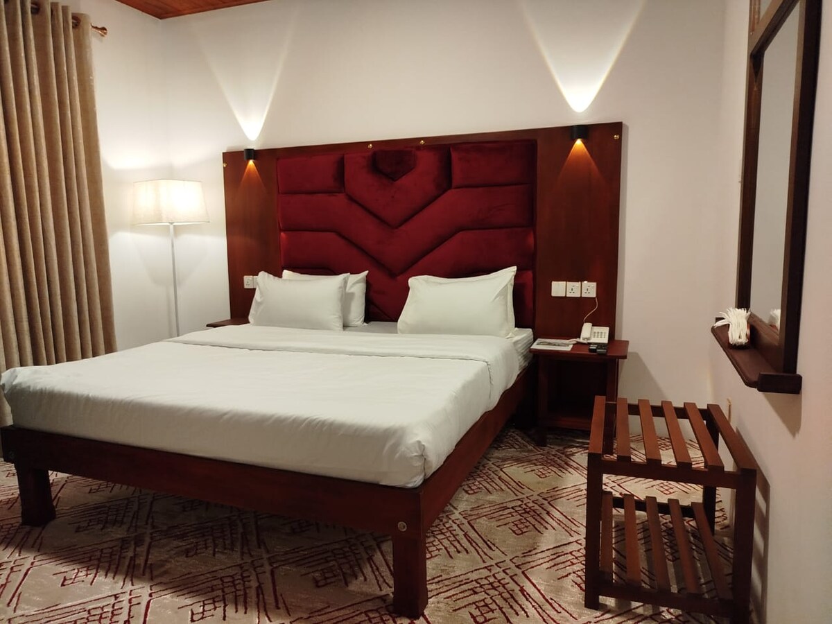 A spacious bedroom features a king-size bed with a red upholstered headboard. Warm lighting is provided by bedside lamps and a standing lamp. A wooden side table and a small nightstand are present. Curtains frame the window, and a soft rug adds texture to the floor.