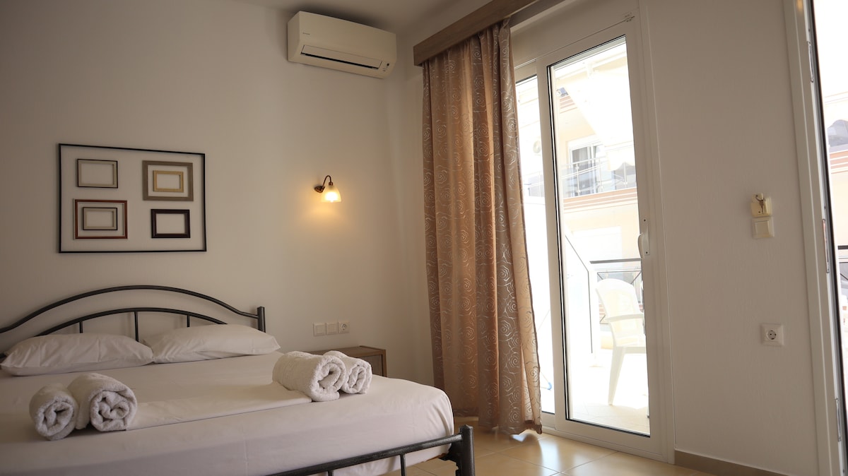 A bright bedroom features a spacious bed with neatly arranged towels at the foot. A sliding glass door opens to a balcony, allowing natural light to fill the room. The soft beige curtains complement the light-colored walls, enhancing the overall calm atmosphere.