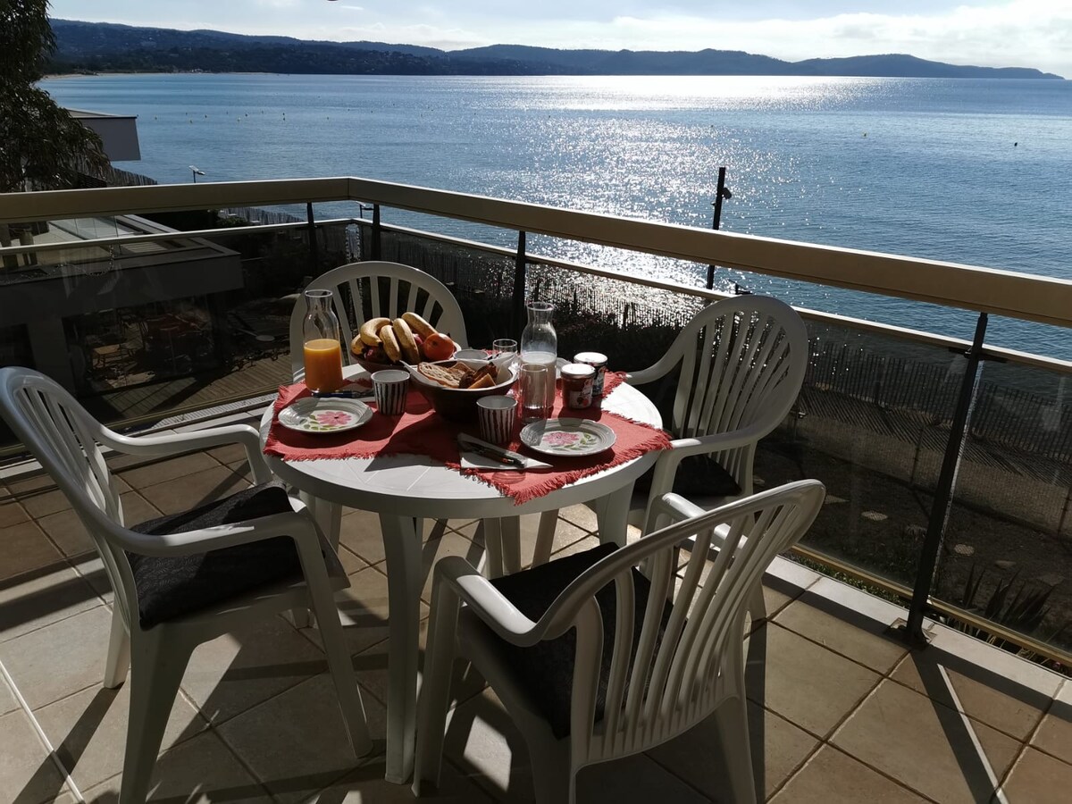 A dining table is set for a meal on the terrace, featuring a colorful tablecloth. Four white chairs surround the table, which holds beverages and food items. In the background, a shimmering body of water reflects light, framed by distant hills under a clear sky.