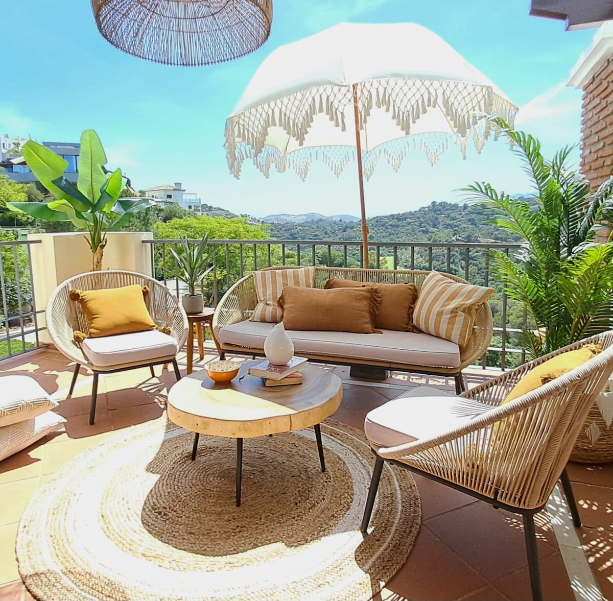 An inviting outdoor terrace is furnished with a cozy seating area, featuring a round table and comfortable armchairs. A large, stylish umbrella provides shade, while lush greenery and mountains are visible in the background under a clear blue sky.