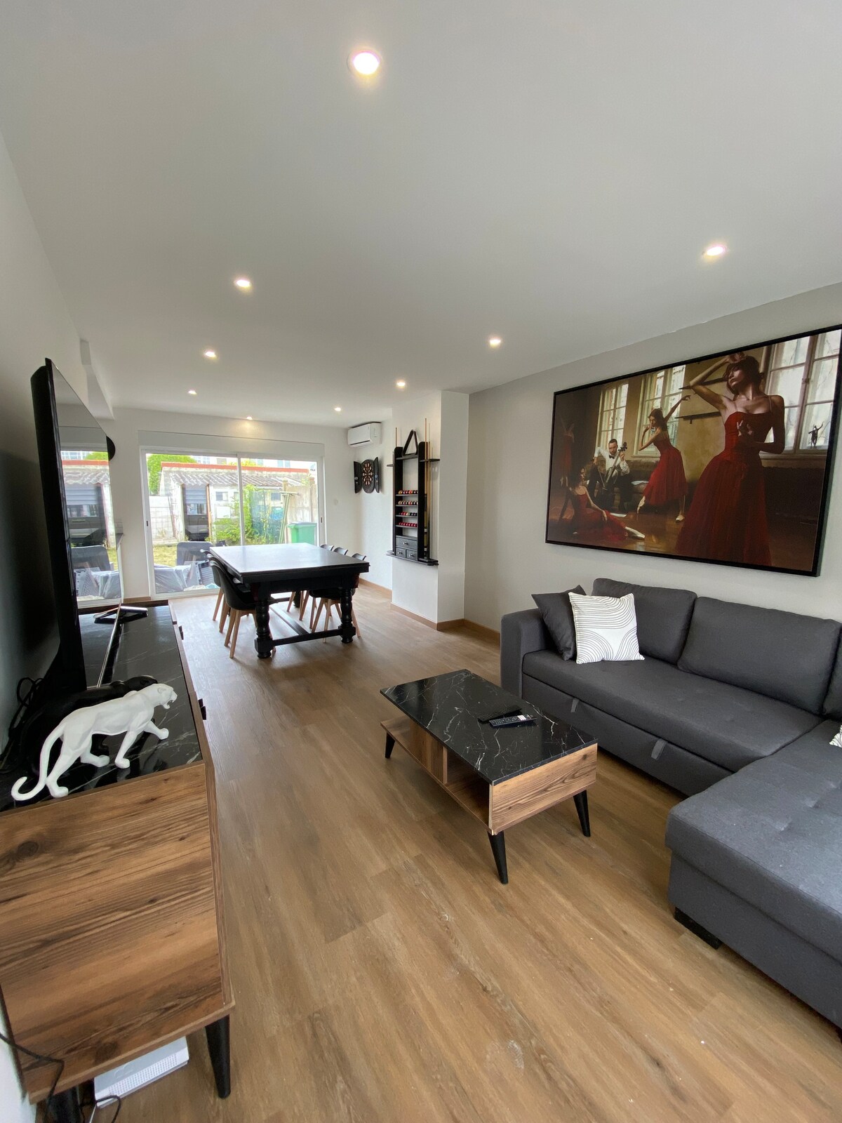 The living area features a spacious layout with elegant wooden flooring. A comfortable gray sofa is positioned beside a sleek coffee table. A large flat-screen TV is mounted on the wall, while a stylish painting adds visual interest. A billiard table is visible in the background.