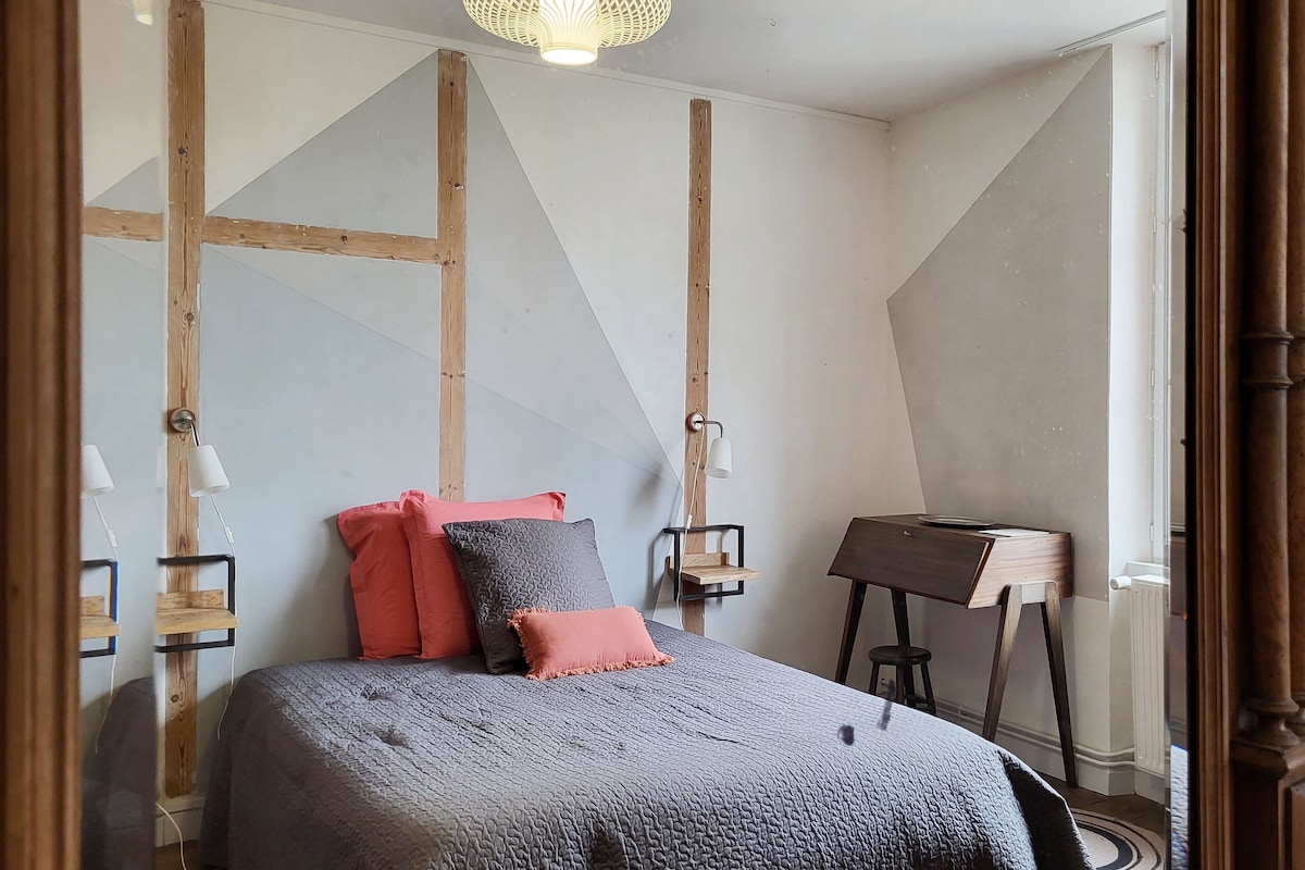 A modern bedroom features a spacious bed with grey bedding and contrasting coral cushions. Simple wooden side tables with bedside lamps are positioned on either side. A stylish wooden desk stands against the wall, complemented by geometric wall accents and natural light filtering through the window.