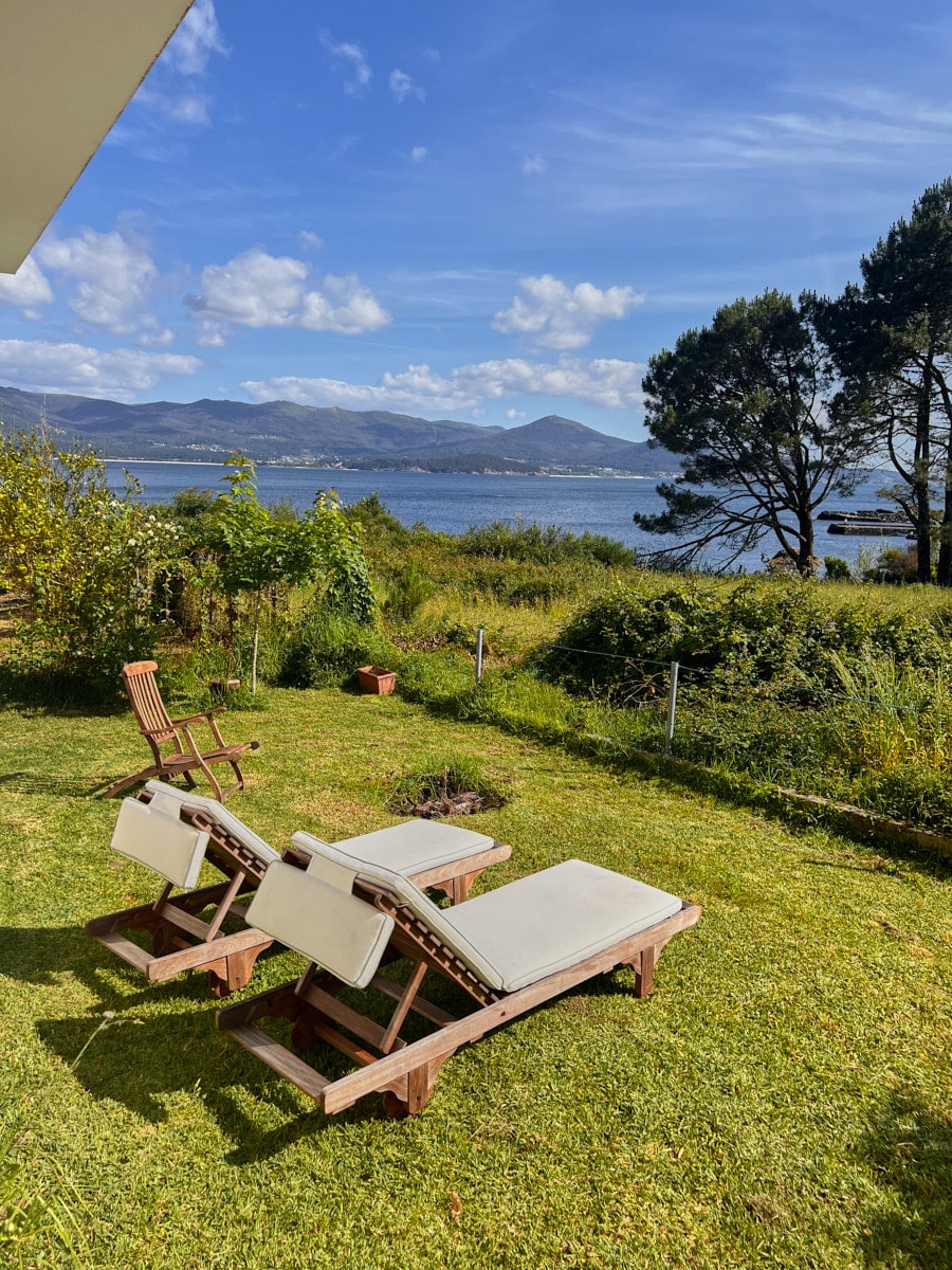 Two comfortable lounge chairs are positioned on a lush green lawn, facing scenic views of the water and distant mountains. The landscape is accented by trees and shrubbery, and the sky is filled with scattered clouds, enhancing the serene outdoor setting.
