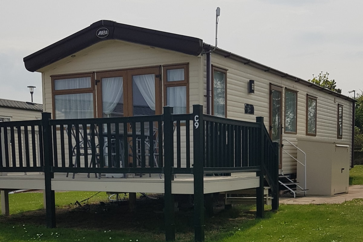 A mobile home is featured, elevated on a grassy area. The exterior includes a wraparound deck with a railing and seating arrangement. Large windows with sheer curtains allow natural light inside. A set of steps leads to the entrance, while the surrounding landscape offers open space.